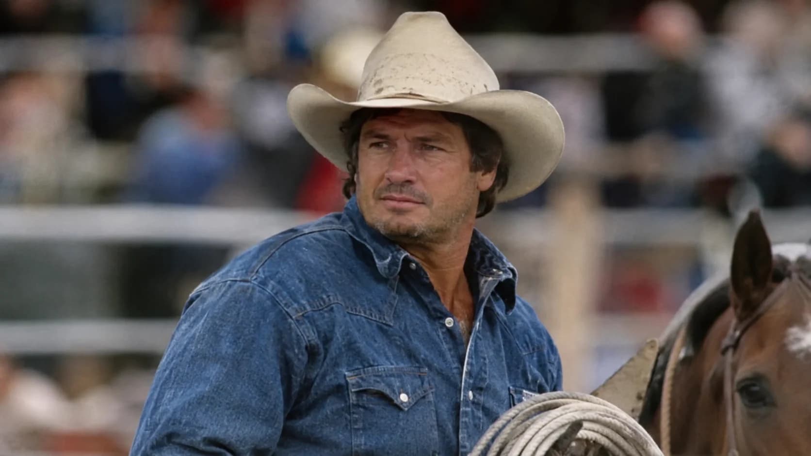 Charlie Battles, American rodeo steer wrestler and rancher, wearing a cowboy hat and denim shirt at a rodeo arena.